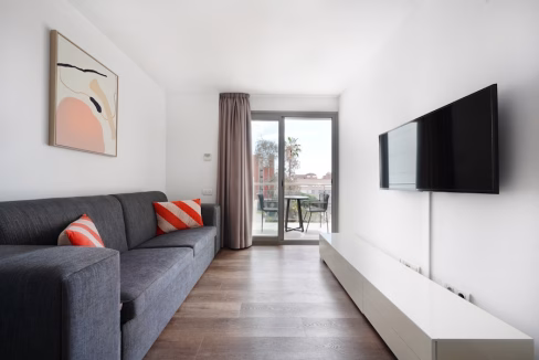 A modern living room featuring a gray sofa with orange and white cushions and a low white entertainment unit. A flat-screen TV is mounted on the wall opposite a large glass door leading to a balcony with outdoor seating.