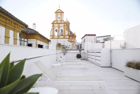 A rooftop terrace featuring white lounge chairs and planters with greenery. In the background, a striking yellow and orange church tower is visible against a light sky. This image showcases the outdoor space at Casa de Salinas Apartments.