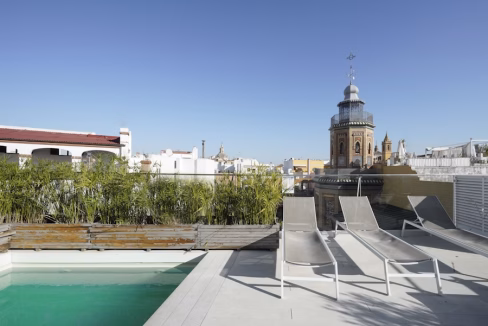 A rooftop area featuring a swimming pool, gray lounge chairs, and potted greenery, with a view of La Giralda Apartments in the background against a clear blue sky. The scene highlights urban architecture and outdoor relaxation spaces.