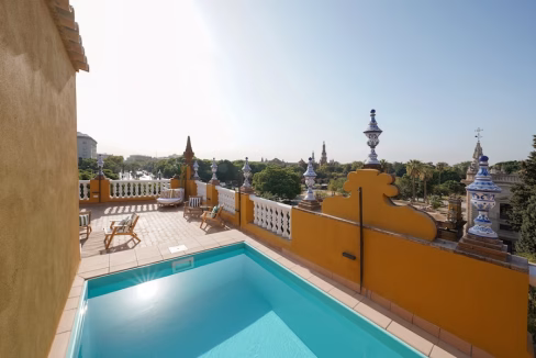 A rooftop terrace with a small swimming pool surrounded by a yellow wall and decorative tiles. The area features lounge chairs and offers a view of lush greenery and distant architectural structures under a clear sky at Murillo Gardens Apartments.