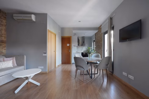 A modern, light-filled living area in the Carrer dels Carders Apartments features a gray wall, a beige sofa with decorative pillows, a white coffee table, and a dining area with four gray chairs. An open kitchen is visible in the background.