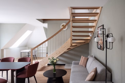A modern interior of the Langstrasse Stauffacher Apartments showcasing a wooden staircase, seating area with a gray sofa, and a round black table surrounded by burgundy chairs. Natural light illuminates the space through a window.
