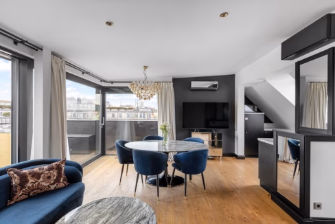 A modern living area in the St. Peter’s Quarter Apartments featuring a blue sofa, a round dining table with blue chairs, and a chandelier. Large windows allow natural light, and a television is mounted on the wall opposite the seating area.