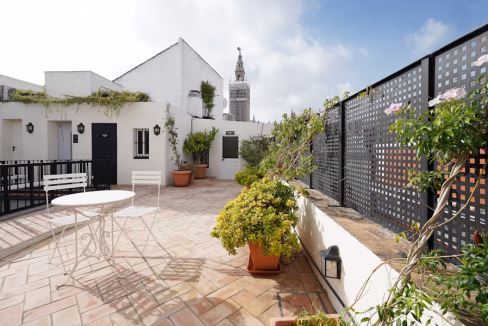 A rooftop terrace featuring a round table and two chairs surrounded by potted plants. In the background, the tower of Seville Cathedral is visible, framed by a clear blue sky. This setting is part of the Seville Cathedral Apartments.