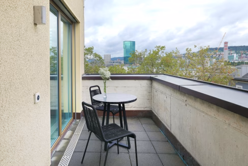 A small balcony featuring a round table with a vase of flowers and two black chairs. The railing provides a view of greenery and distant buildings under a cloudy sky, highlighting the outdoor space of Langstrasse Schoneggstrasse Apartments.