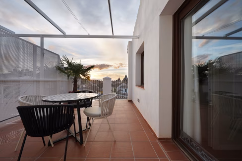 A balcony area with a round table and four chairs, two are black and one is white. A potted palm tree is present, and the background features the skyline under a sunset with colorful clouds. This is part of the Granada Gran Via Apartments.