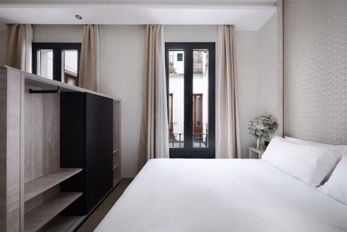A minimalist bedroom featuring a white bed with crisp linens, a black and wooden wardrobe, and large windows with sheer curtains. A small vase with flowers sits on a nearby table, while light fills the space from the windows.