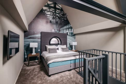A cozy bedroom featuring a large bed with gray and white bedding, flanked by two bedside tables with lamps. The wall behind the bed displays a mural of a warehouse interior, and the room has a sloped ceiling with modern angles.