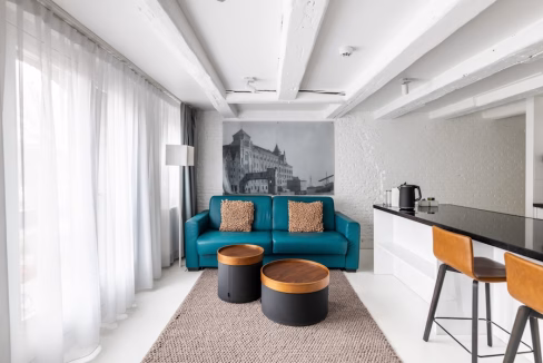 A modern living area featuring a teal sofa with two decorative pillows, two round coffee tables, and a patterned rug. In the background, there's a sleek kitchen counter with bar stools and a large window letting in natural light. This decor is part of the Salthouse Canal Apartments.