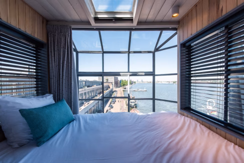 A view from inside a bedroom with a bed covered in white linens and a teal pillow, featuring large windows displaying Amsterdam Harbour with boats and waterfront pathways. The warm wooden interior and window blinds are also visible.