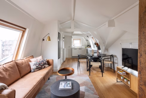 A stylish living area in the Vondelbunker Apartments features a brown sofa with decorative pillows, a round black coffee table, and a dining set with four black chairs. Natural light streams through a slanted window, illuminating the wooden floor and modern decor.