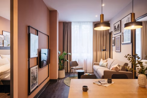 A modern living space featuring a light brown sofa, a decorative area rug, and a wall-mounted television. The room has large windows with sheer curtains, potted plants, and framed artwork on the walls, creating a cozy atmosphere in the The Hague Willemspark Apartments.