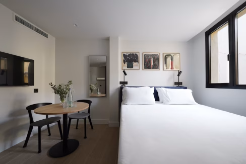 A modern bedroom in the Madrid Sol Apartments featuring a double bed with white linens, two black chairs, and a round wooden table. The room has clean lines, natural light from large windows, and artwork on the wall.