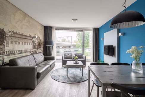 A modern living area featuring a gray leather sofa, a round rug, and a black coffee table with a small plant on it. The wall behind has a mural of buildings, while a blue wall displays a flat-screen TV. Large windows allow natural light to enter.