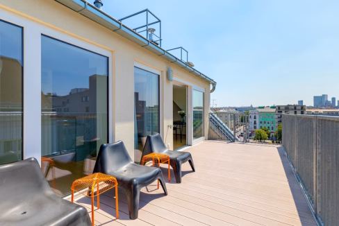 A spacious rooftop terrace with wooden flooring features three black plastic chairs and two orange side tables. Large glass doors lead into the interior, while cityscape views are visible in the background. This setting is part of the Vienna Stiegergasse Apartments.
