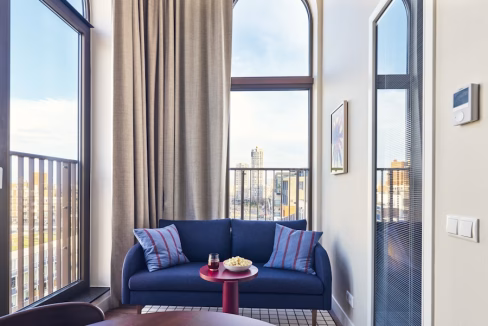 A cozy sitting area featuring a blue couch with striped cushions beside a round red table. Large windows allow natural light to fill the space, with sheer curtains and a city view visible outside. This setting is part of The Hague Beach Apartments.
