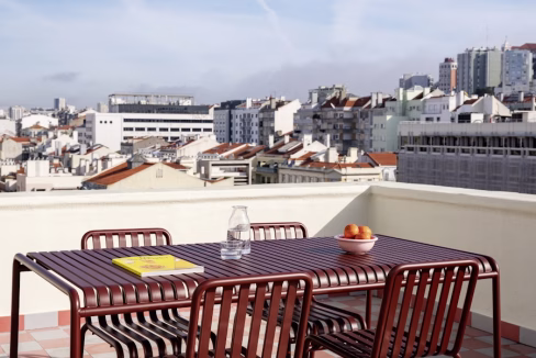A rooftop terrace with a metal dining table and six chairs, set with a glass pitcher and a bowl of oranges. The background features a cityscape of buildings and rooftops under a clear sky, representing the Santa Cruz Anjos Apartments.