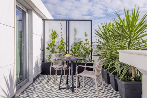 A balcony with a tiled floor features a round black table surrounded by two light-colored chairs. Potted plants line the space, and a backdrop of greenery is visible behind a privacy screen. This is part of the Principe Real Rato Apartments.