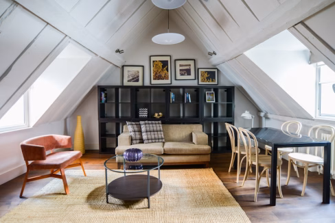An attic apartment furnished with a beige sofa, an orange armchair, and a black dining table with wooden chairs. A round coffee table sits on a woven rug, and framed artworks are displayed on the wall above a black bookshelf. Light filters through the windows.