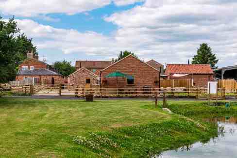 Farm buildings at York Brecks Farm with lush green fields and a pond under a partly cloudy sky.