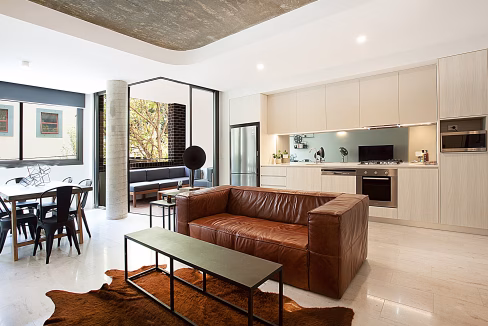 Interior view of a modern living space at Belmore Park Apartments, featuring a brown leather couch, a coffee table, and a dining area with a black table. The kitchen visible in the background includes stainless steel appliances and cabinetry.