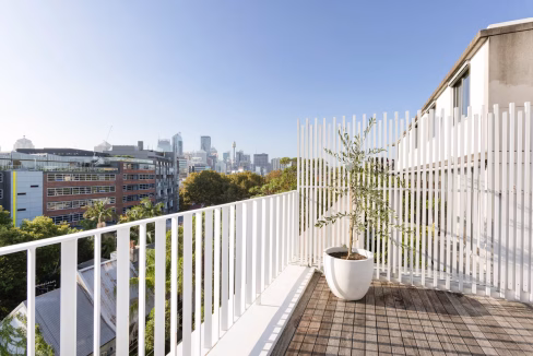 A balcony with a potted plant overlooks a cityscape with various buildings in Sydney. The white railing creates a modern feel, framed by clear skies. This setting is part of the Sydney Central Station Apartments, highlighting urban living.
