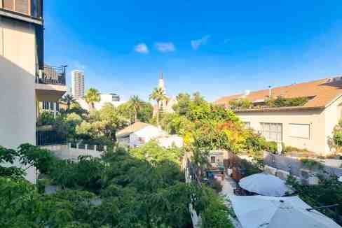 View of Nitsana Street Apartments surrounded by trees and modern buildings under a clear blue sky.