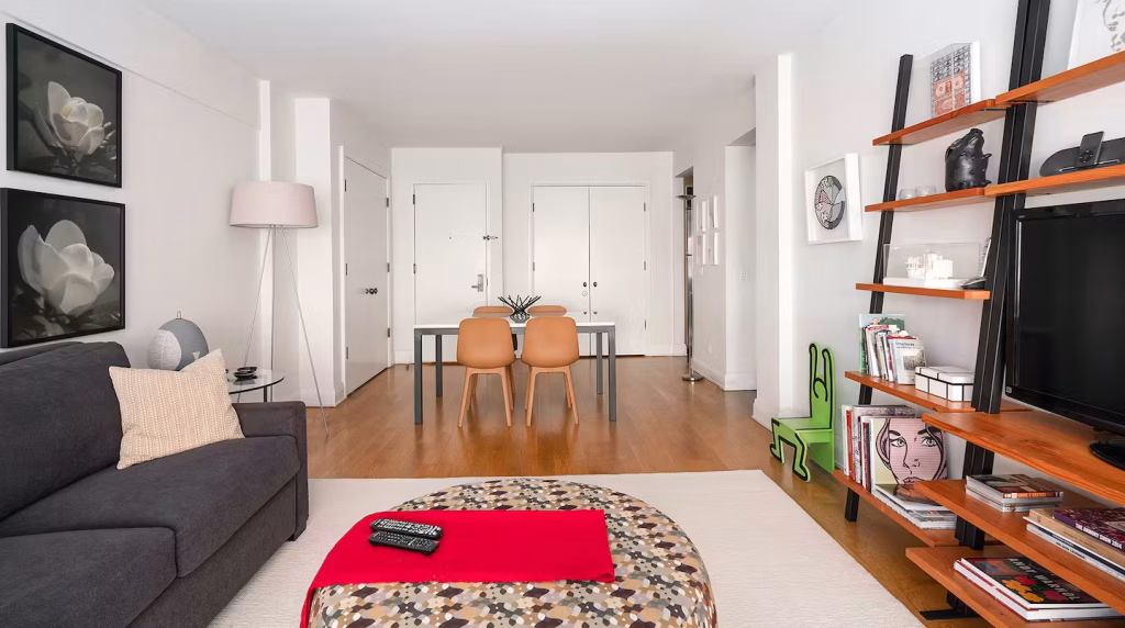 A living room in an apartment featuring a gray sofa, a round patterned coffee table, and a dining area with two orange chairs. The space includes light wood flooring and white walls, with artwork hanging on the walls, reflecting a modern design in Manhattan Midtown East Apartments.