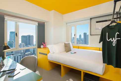 Modern hotel room with large windows overlooking Manila skyline. Bright yellow ceiling and cozy bed.