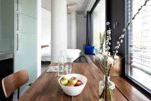 Bright city apartment with dining table, fresh fruit, and decorative flowers by large window.
