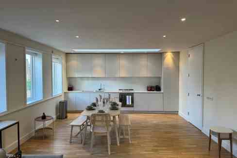 Modern minimalist kitchen and dining space with natural light, white cabinets, and wooden flooring f.
