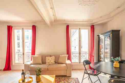 Bright living room featuring large windows with red curtains, a beige sofa, and a black dining table.