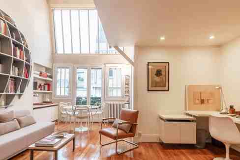 Bright modernist salon with large windows, bookshelf, and cozy seating.