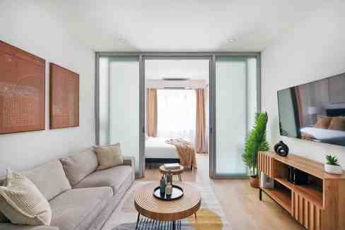 Spacious modern living room with a cozy sofa, wooden coffee table, and a view into a bright bedroom.