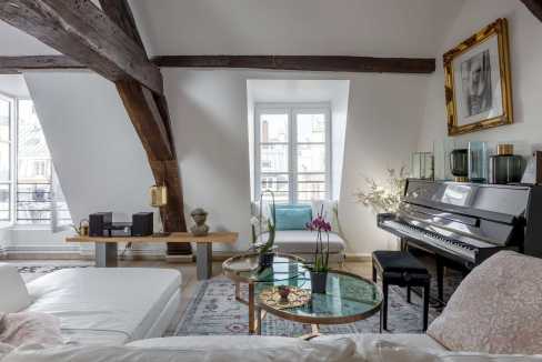 A cozy living room with a grand piano, exposed wooden beams, and large windows letting in natural li.
