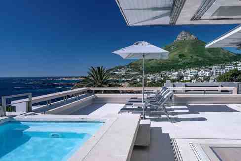 Seaside terrace with pool, sun loungers, and mountain backdrop in a luxury setting.