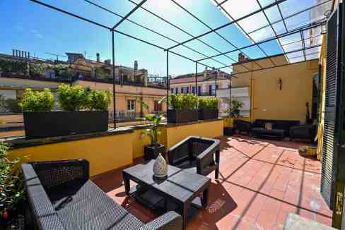 Rooftop terrace with outdoor furniture, potted plants, and glass roof in an urban setting.