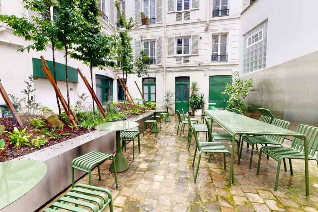 Charming outdoor courtyard with green tables and chairs, surrounded by lush plants and urban archite.
