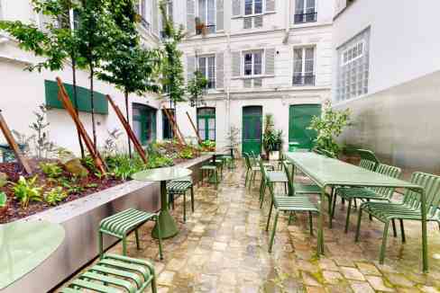 Charming outdoor courtyard with green tables and chairs, surrounded by lush plants and urban archite.