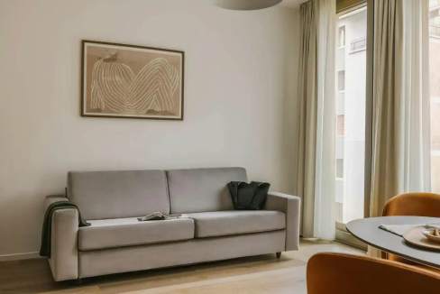 A gray sofa with a black bag resting on it sits against a light-colored wall. Above the sofa, an abstract art piece is framed, and floor-to-ceiling curtains allow natural light. A round table and chairs are visible in the background, contributing to the Montenero Urban Luxe aesthetic.