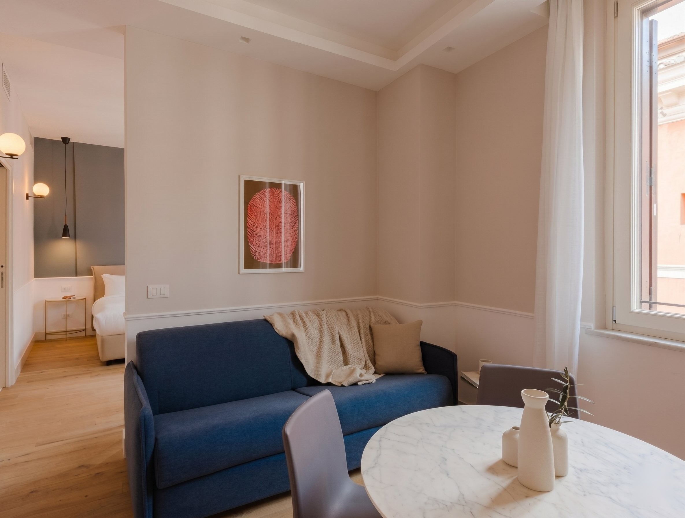A cozy room featuring a blue sofa with a beige throw, a round marble table with a white vase, and a framed artwork on the wall. Natural light enters through a large window, illuminating the neutral-toned walls. This setting reflects the Condotti Roma Elegance.