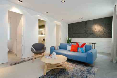 A modern living room in Chueca Rhythm Retreat with a blue sofa, orange pillows, a round wooden coffee table, and recessed ceiling lights beside white walls and open doorways.