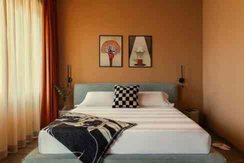 A modern bedroom in the Milan Ticinese Loft, featuring a bed with a checkered black and white pillow and a cozy blanket. Two framed artworks are on the rust-colored walls, with sheer curtains allowing natural light to illuminate the space.
