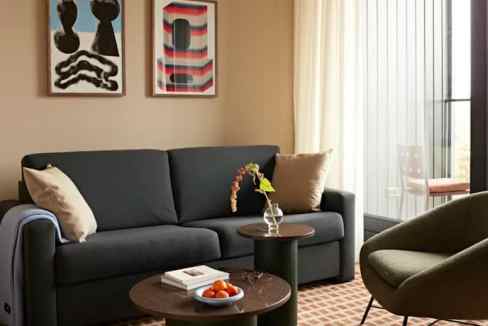 A modern living area featuring a dark gray sofa with two pillows, flanked by two circular tables. One table holds a small vase with a plant and a stack of books. The space has art on the walls and large windows allowing natural light, part of Vondelpark City Suites.