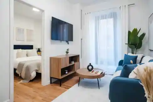Bright living room with blue sofa, wall-mounted TV, and large window with white curtains.