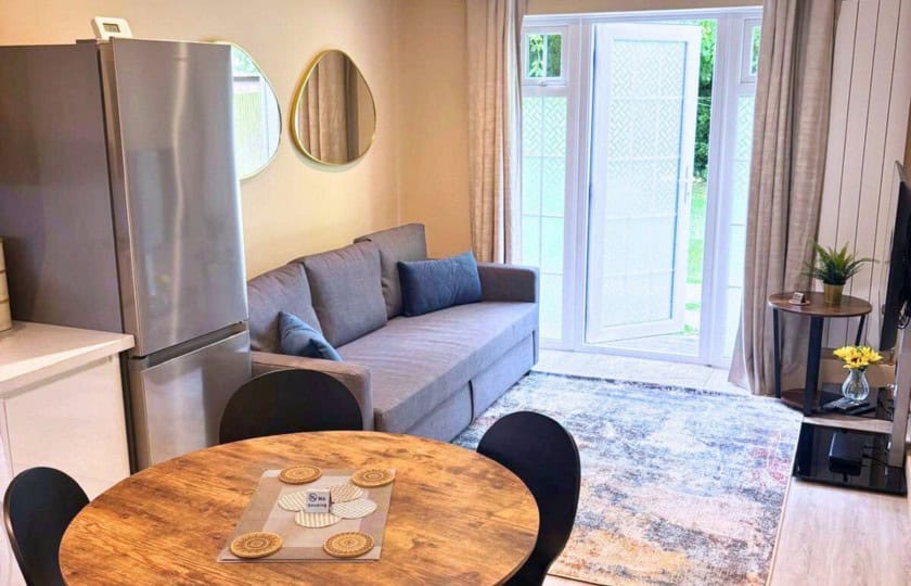 A compact living room in the Colindale Yellowhammer Suite with a gray sofa, round wooden dining table, and stainless steel refrigerator beside glass doors and a patterned rug.