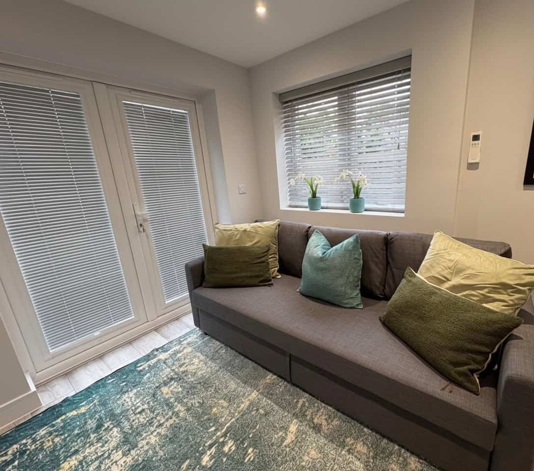 A gray sofa with green and blue cushions sits in a bright room with blinds on the doors and window, a patterned rug below, and Glenthorne New Southgate in the room context.