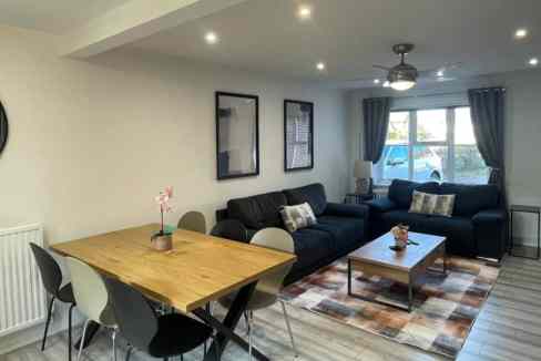 A modern living room at Hayes House Sanctuary with two dark sofas, a coffee table on a patterned rug, and a wooden dining table with chairs beside a window with curtains.