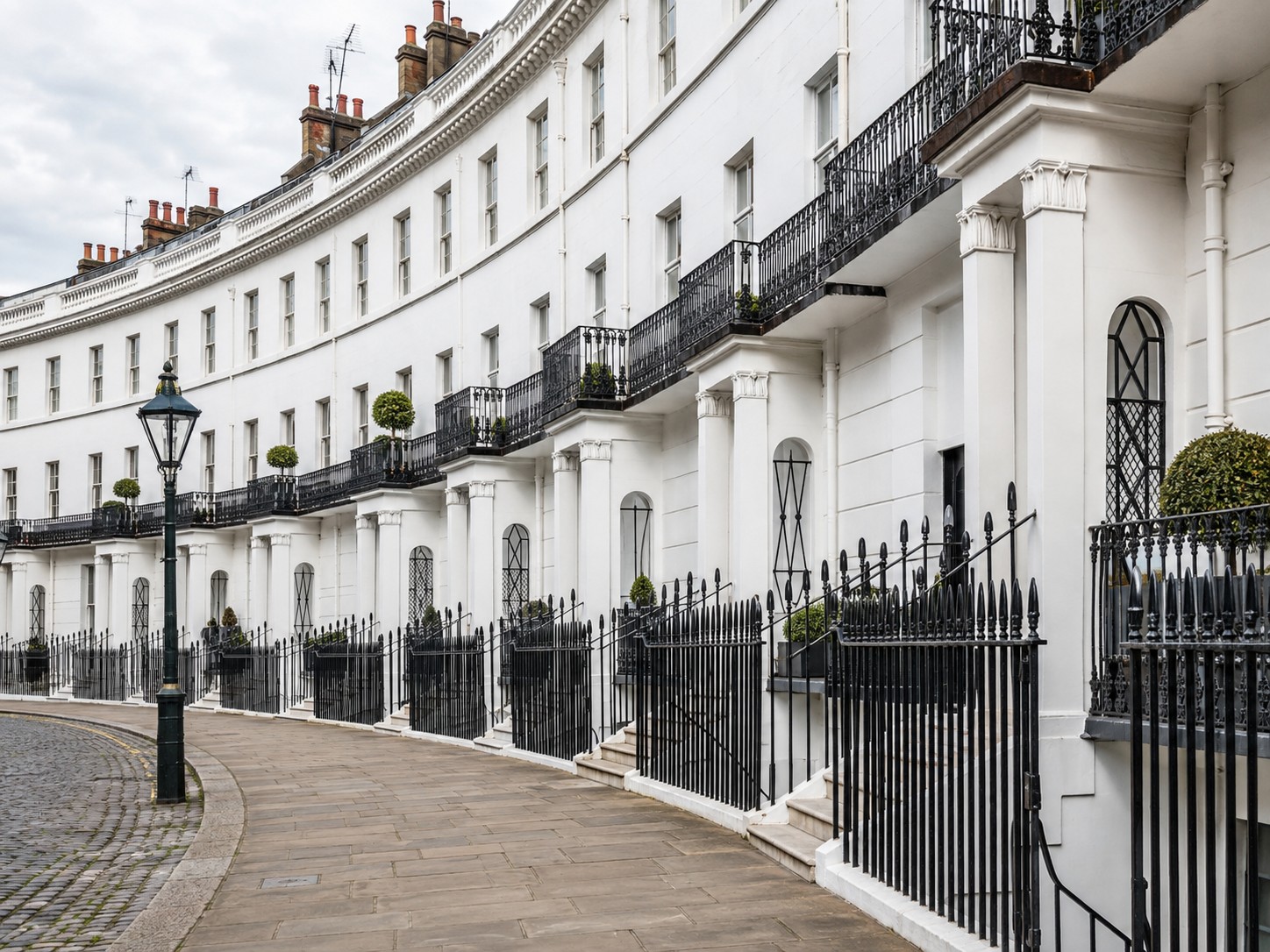 A curved row of white Georgian townhouses with black wrought-iron balconies and railings along a cobblestone street in South Kensignton