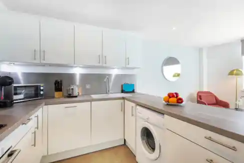 A bright modern kitchen at Camden Regent Retreat with white cabinets, wood countertops, and a washer under the peninsula, plus a bowl of fruit and a round wall mirror.